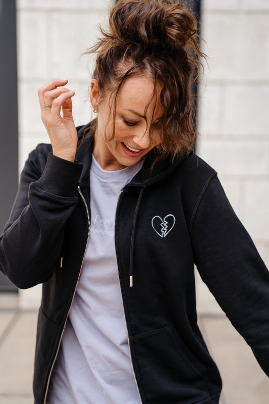 Vorne geöffneter schwarzer Zip-Hoodie mit kleiner, weißer Herz-Stickerei auf der Brust. Darunter sichtbar: ein pastellfarbenes T-Shirt mit weißem „perfectly imperfect“-Print.