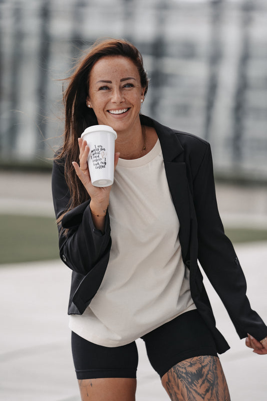 Weiße Mehrweg-Kaffeetasse mit Deckel, in beiden Händen vor dem Oberkörper gehalten. Der schwarze Aufdruck auf der Vorderseite lautet: „I am sorry for what I said before I had my coffee“, ergänzt durch ein kleines gezeichnetes Herz in gold-beiger Farbe. Die Tasse wird von einer Person mit hellem Oberteil und dunklem Blazer präsentiert.