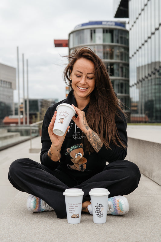 Weiße Trinkbecher-Tasse mit Deckel und Aufdruck „drinking buddy!“ in brauner Schreibschrift. Darunter ist ein niedlicher, sitzender Bär mit grünem Stirnband abgebildet.