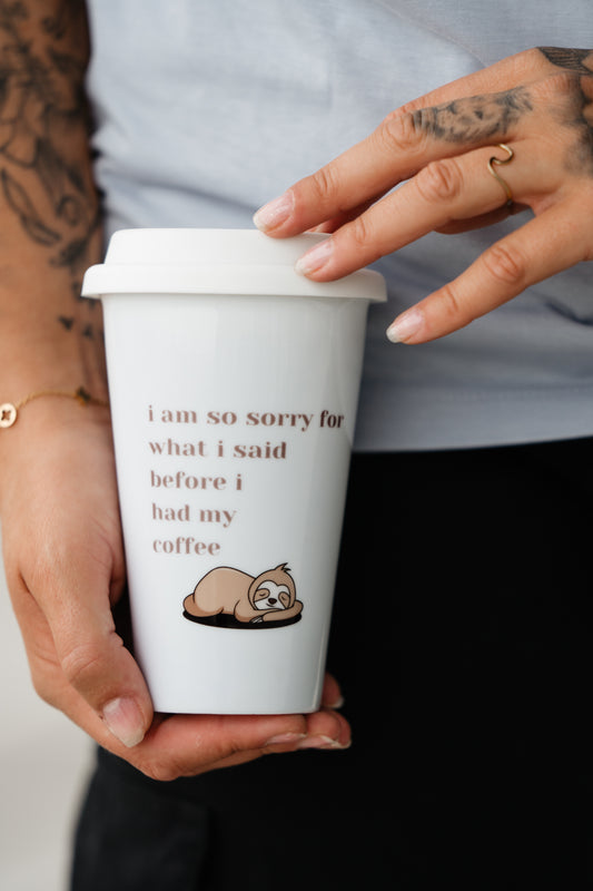 Weiße Mehrweg-Kaffeetasse mit Deckel, in der Hand gehalten. Auf der Vorderseite befindet sich eine Illustration eines schlafenden Faultiers sowie der Aufdruck „i am so sorry for what i said before i had my coffee“ in brauner Schrift. Der Becher ist leicht geneigt, der Deckel wird mit der anderen Hand angedeutet berührt.