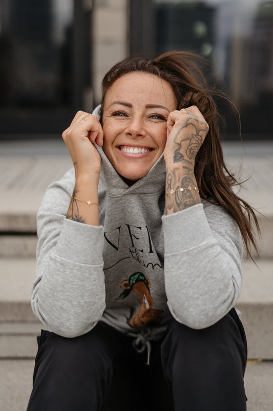 Hellgrauer Kapuzenpullover mit schwarzem Print „OVERthinking“ und einer großen Illustration eines nachdenklich blickenden Teddybären mit grünem Bandana. Der Bär sitzt in der Mitte des Brustbereichs, die Typografie kombiniert Druckschrift und Handschrift.