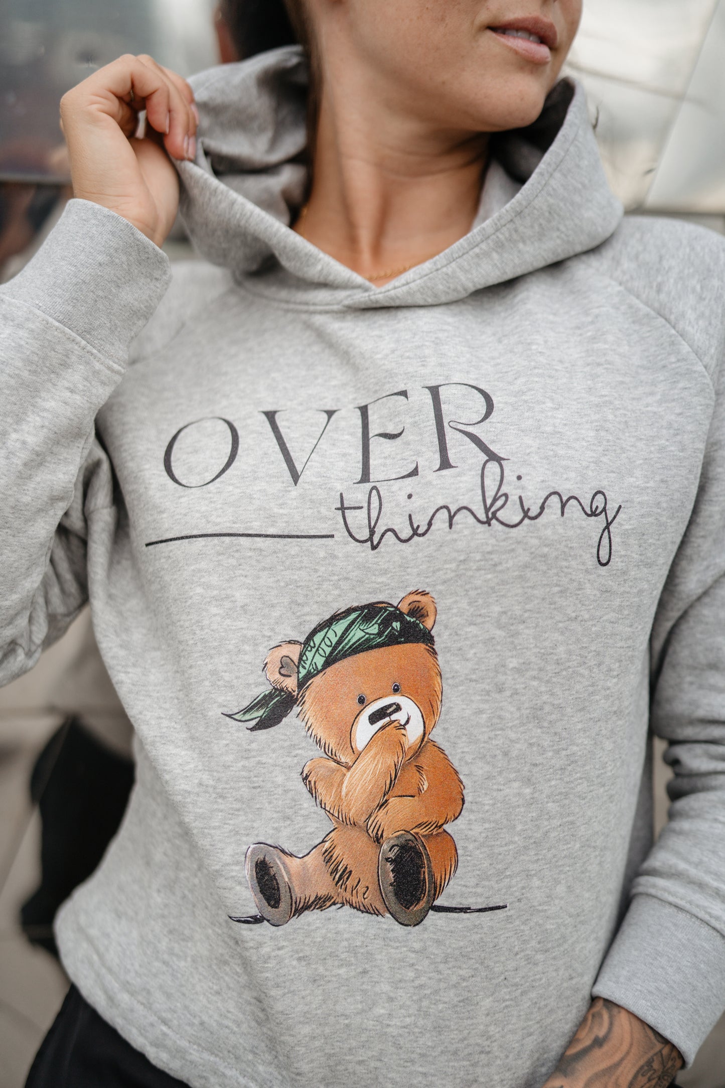 Hellgrauer Kapuzenpullover mit schwarzem Print „OVERthinking“ und einer großen Illustration eines nachdenklich blickenden Teddybären mit grünem Bandana. Der Bär sitzt in der Mitte des Brustbereichs, die Typografie kombiniert Druckschrift und Handschrift.