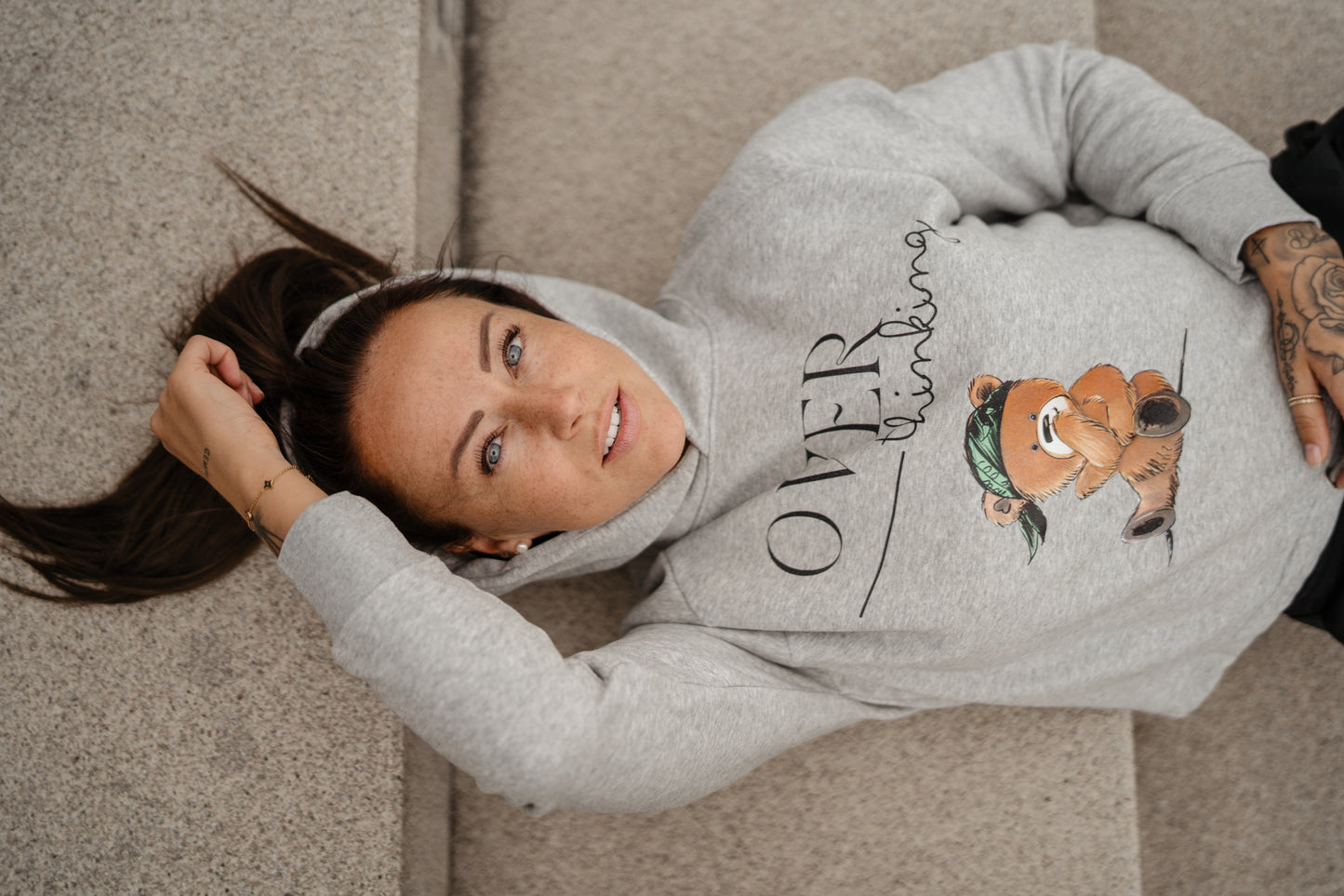 Hellgrauer Kapuzenpullover mit schwarzem Print „OVERthinking“ und einer großen Illustration eines nachdenklich blickenden Teddybären mit grünem Bandana. Der Bär sitzt in der Mitte des Brustbereichs, die Typografie kombiniert Druckschrift und Handschrift.