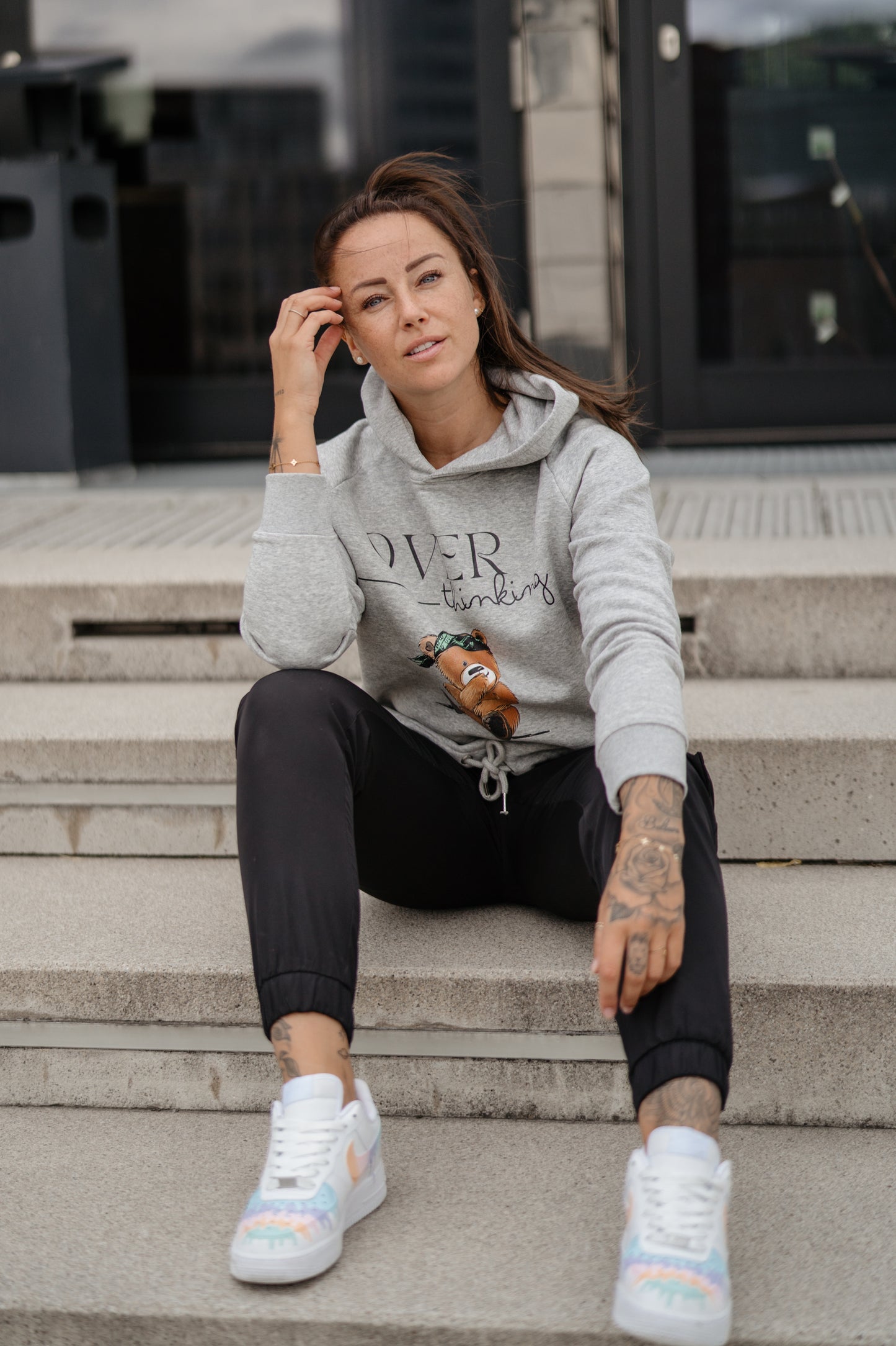 Hellgrauer Kapuzenpullover mit schwarzem Print „OVERthinking“ und einer großen Illustration eines nachdenklich blickenden Teddybären mit grünem Bandana. Der Bär sitzt in der Mitte des Brustbereichs, die Typografie kombiniert Druckschrift und Handschrift.