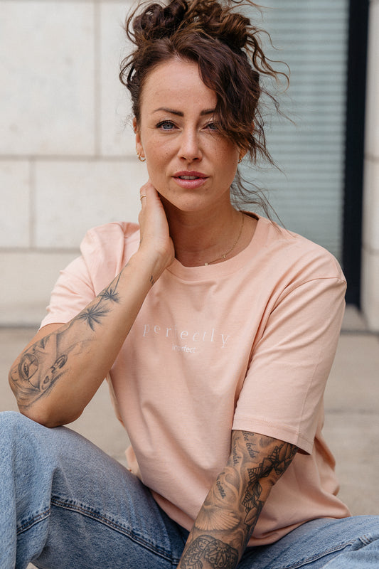 Beigefarbenes T-Shirt mit weißem Frontprint „perfectly imperfect“ in feiner, minimalistischer Typografie. Das T-Shirt besteht aus glattem Baumwollstoff und hat einen klassischen Rundhalsausschnitt sowie eine locker geschnittene Passform.