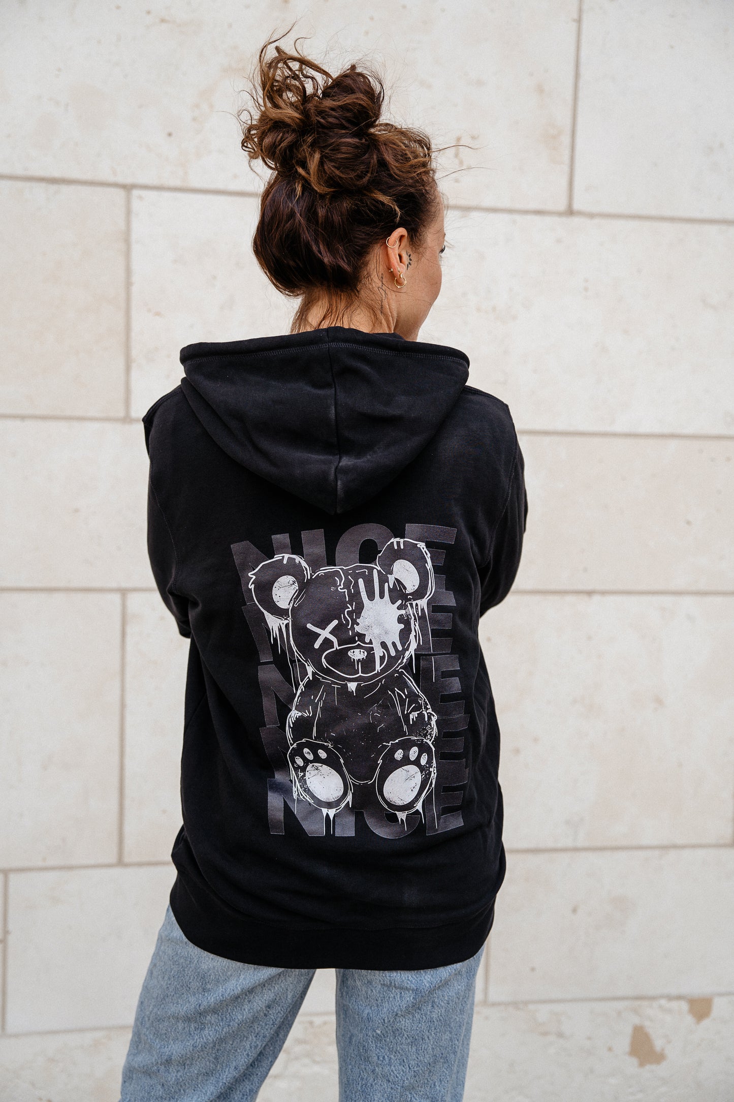 Schwarzer Kapuzenpullover mit Reißverschluss und großflächigem Rückendruck eines stilisierten Teddybären in Grautönen. Der Bär ist mit Spritzer-Optik gestaltet und von dem mehrfach gesetzten Wort „NICE“ hinterlegt.