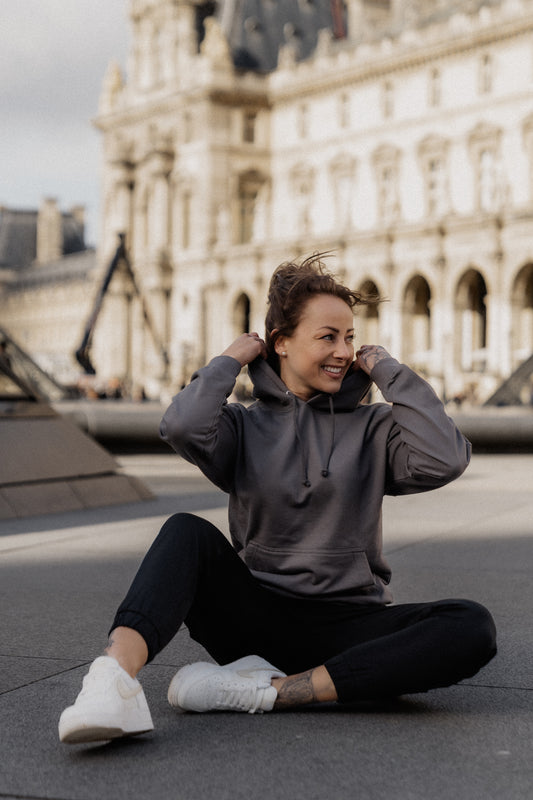Person trägt einen anthrazitfarbenen Hoodie mit großem weißen Rückendruck eines nachdenklich sitzenden Teddybären mit Stirnband und dem Schriftzug „Overthinking…“. Das Sweatshirt wird im urbanen Außenbereich getragen, der Fokus liegt auf dem Design auf dem Rücken.